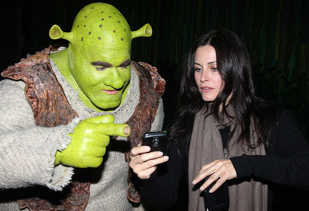 Courtney Cox holding a Blackberry as someone in a Shrek costumes looks on