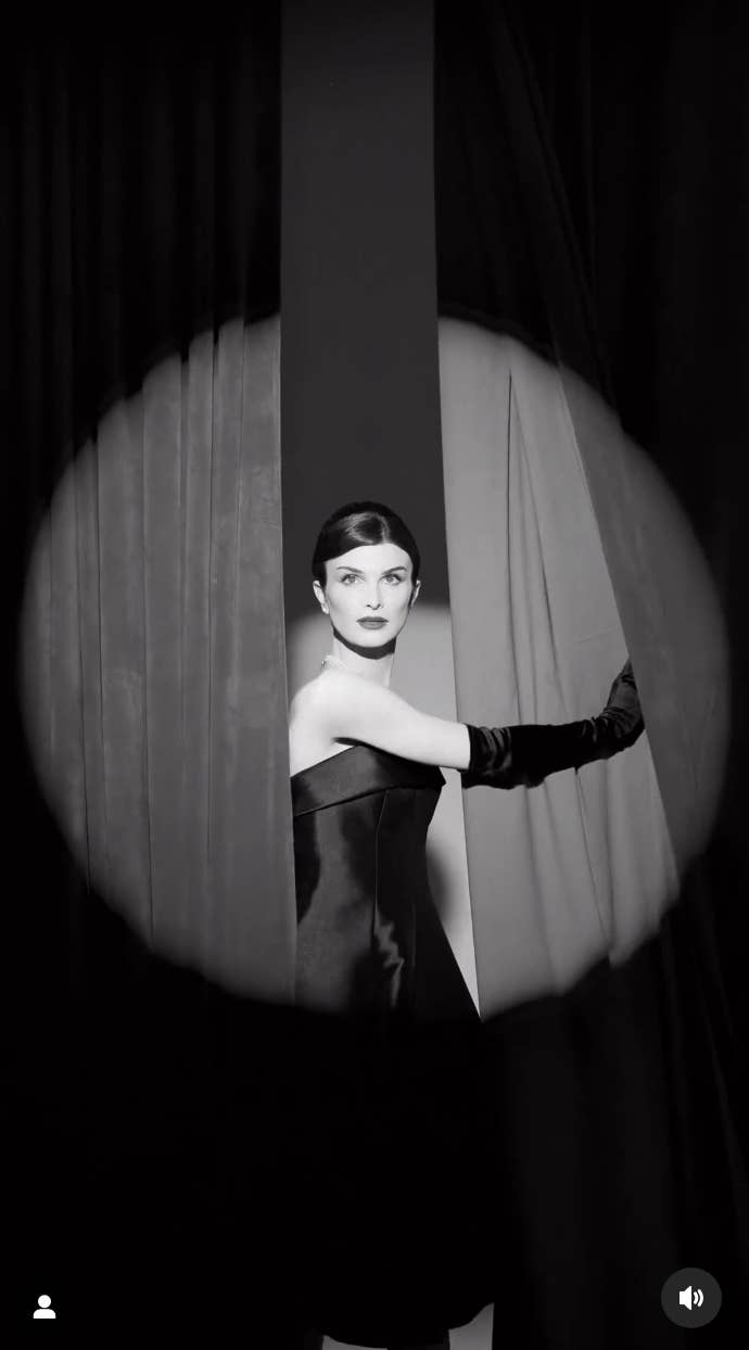 A noir-style black-and-white photo of Dylan standing between two curtains while wearing a satin strapless tea-length dress and matching opera gloves. Dylan, who has a spotlight on them, also has her hair in an updo