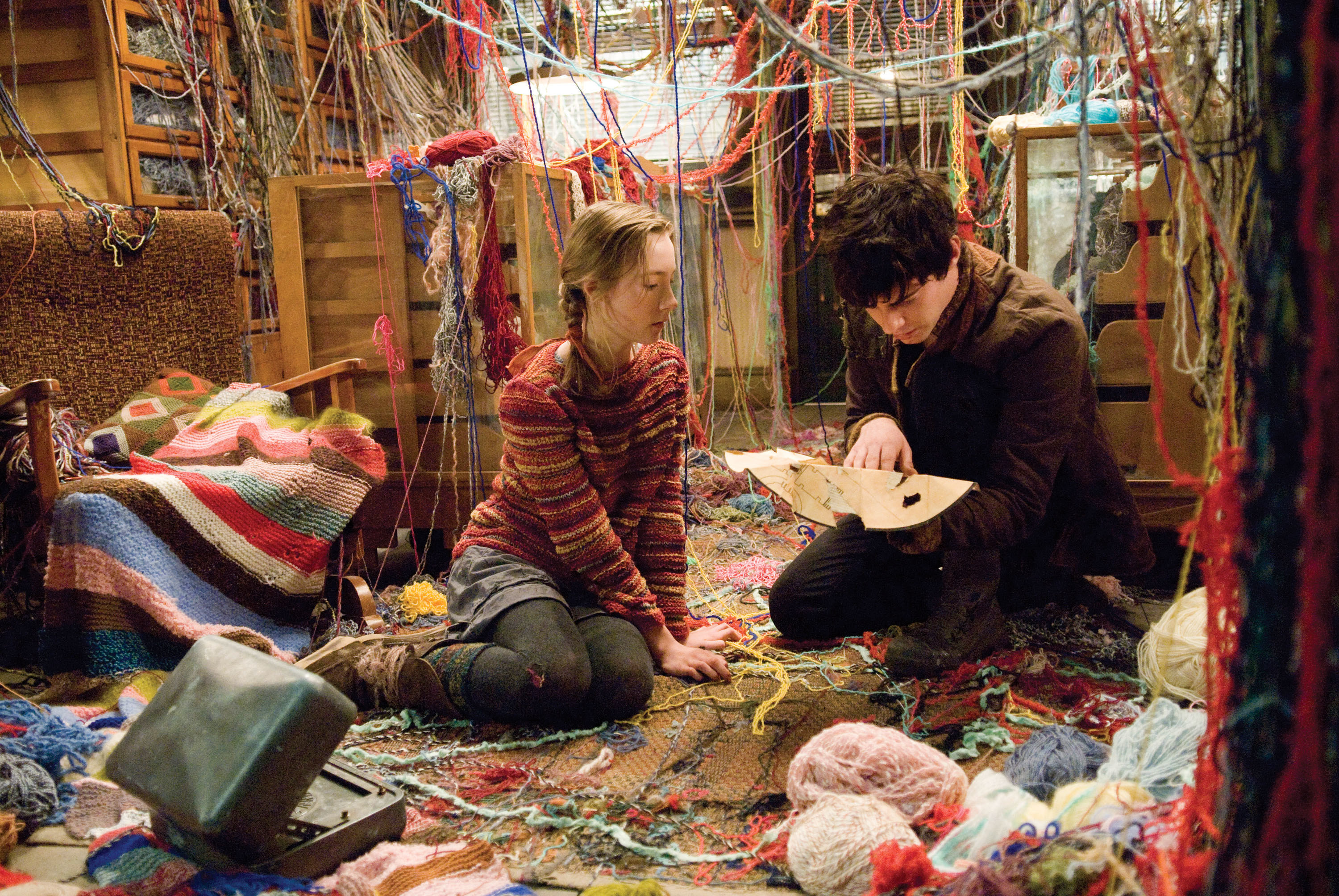 two teens sitting on the floor of a room with yarn everywhere