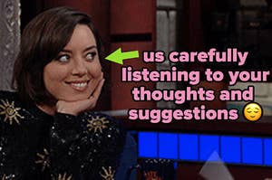 aubrey plaza sits with her hand under her chin, leaning in toward someone off-camera, listening intently