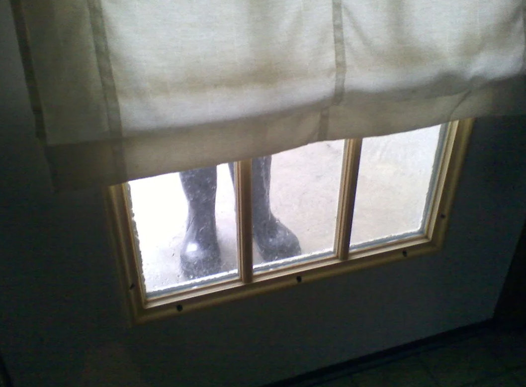 a pair of rubber boots standing just outside a door, and the top part covered by curtains so it looks like someone is standing outside the door