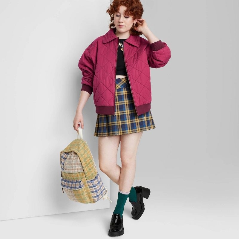 Model wearing pink jacket over black top, blue and yellow skirt and backpack with green socks and black loafers