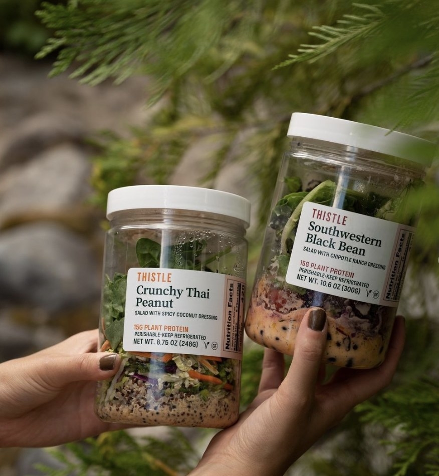 Someone holding jars of crunchy thai peanut salad and southwestern black bean salad in front of fir tree