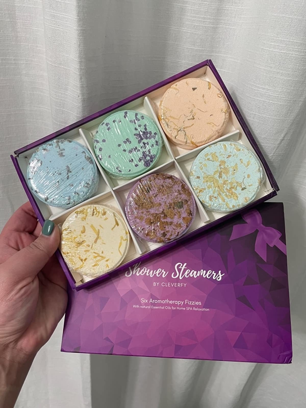 A reviewer holding the box of six shower steamers (blue, green, orange, yellow, purple, mint)