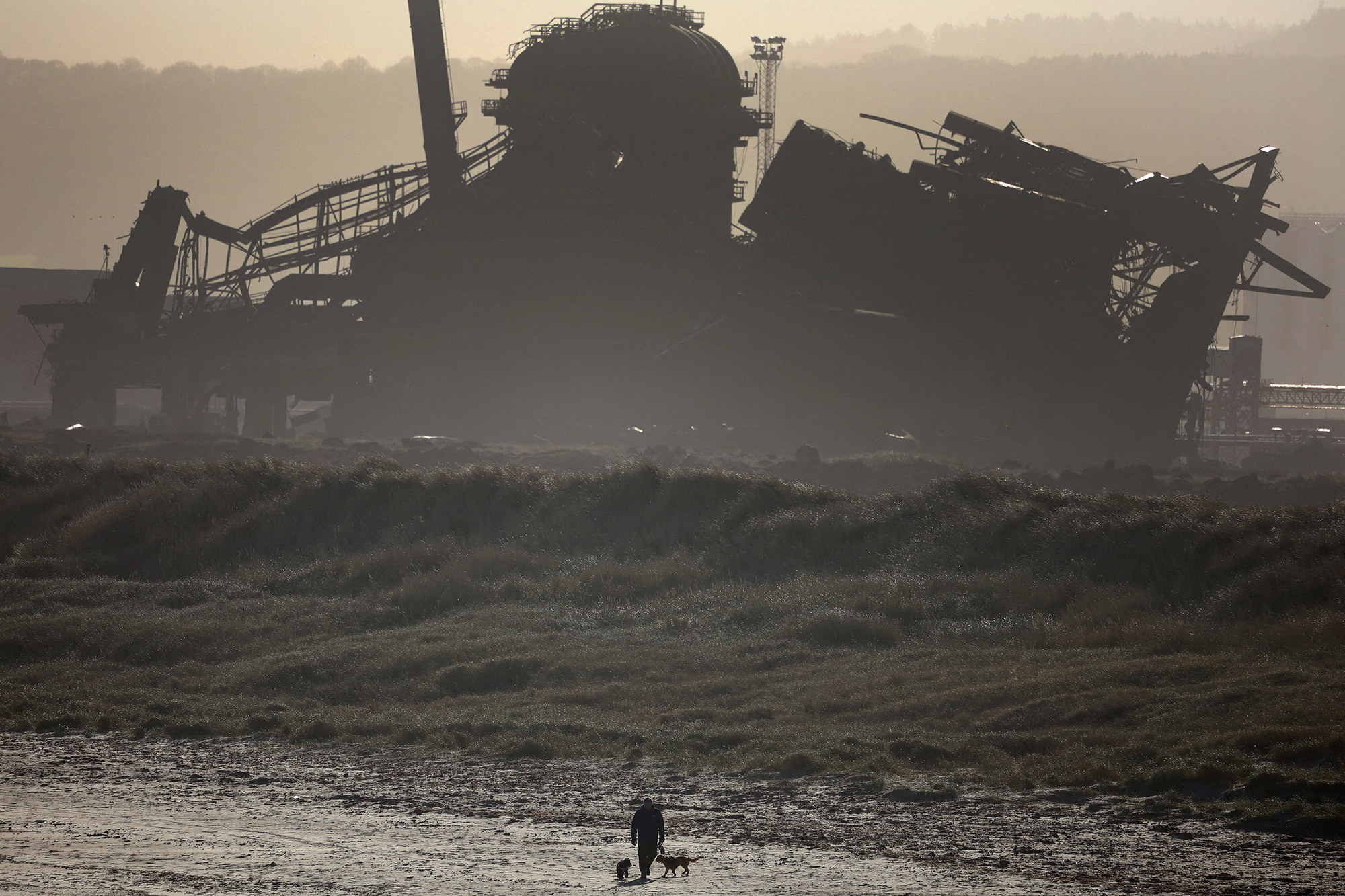 a zoomed out shot of a man walking his dog in front of the shadowy outline of a demolished blast furnace