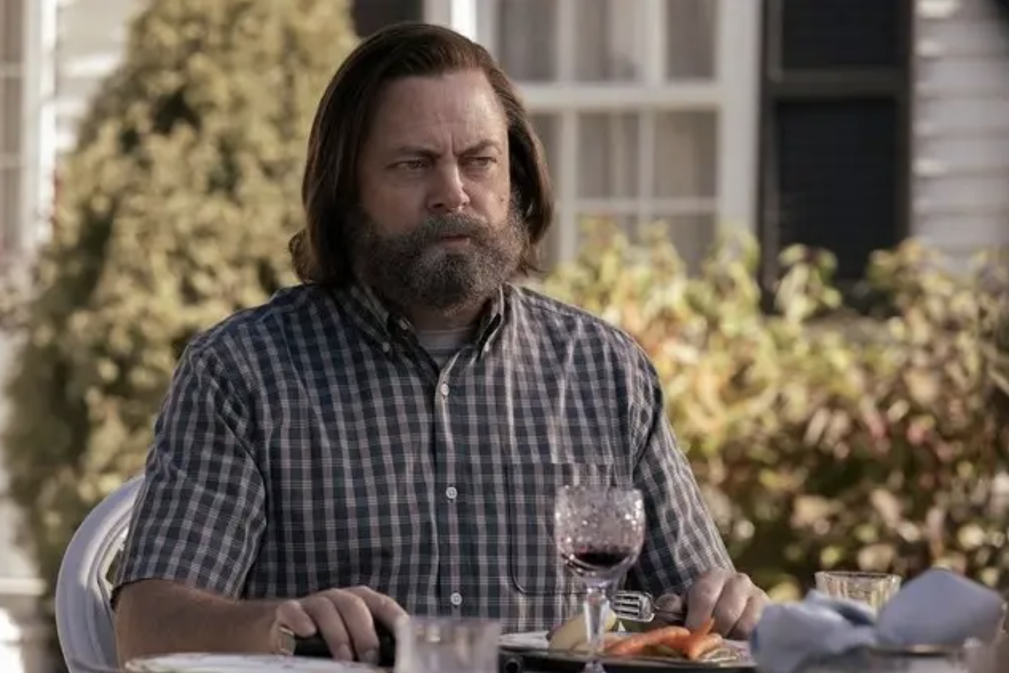 Nick Offerman in a short sleeve button down sitting at a table outside on a sunny day with a glass of wine