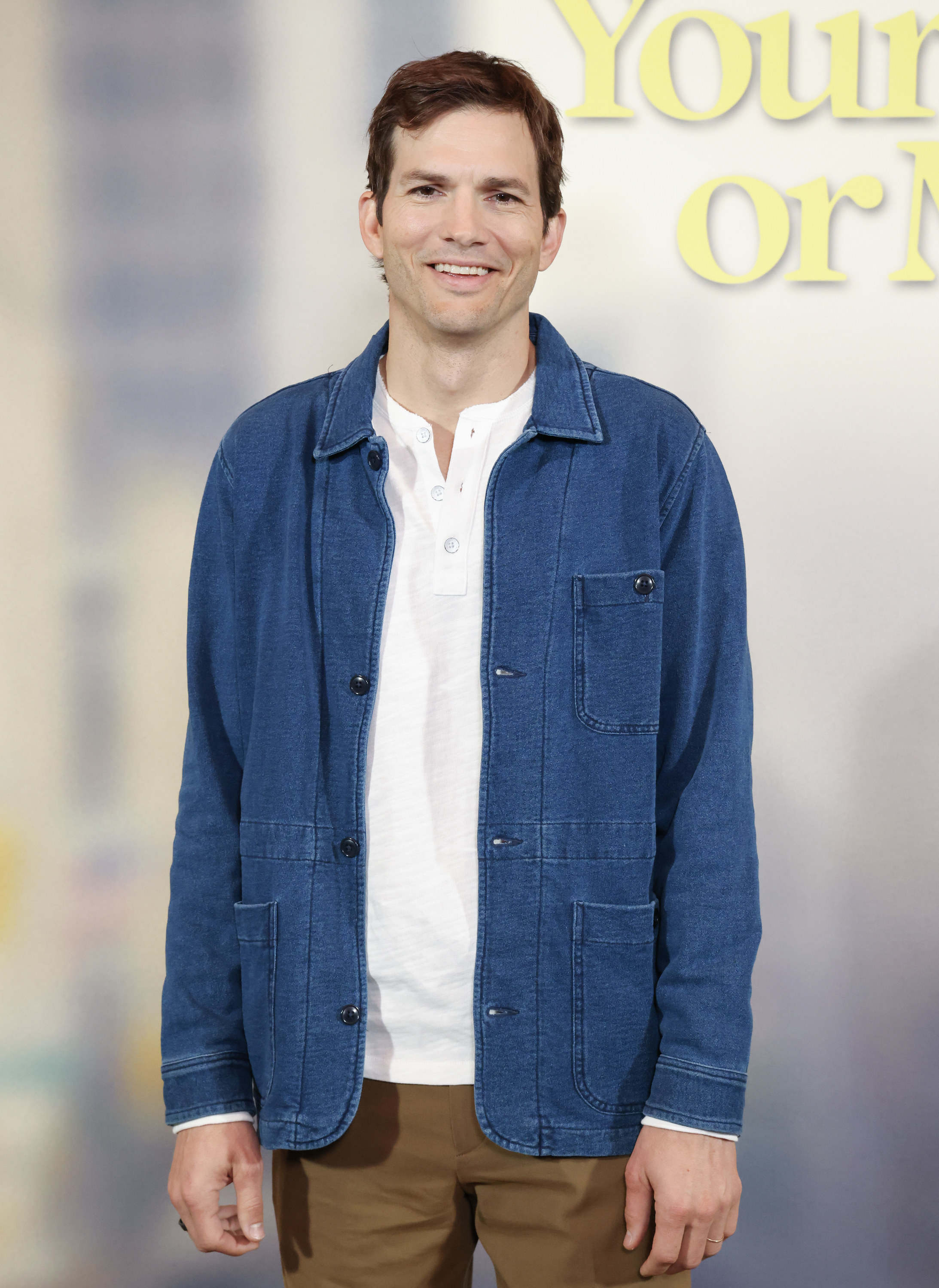 A close-up of Ashton smiling. He&#x27;s wearing a t-shirt, a denim jacket, and slacks