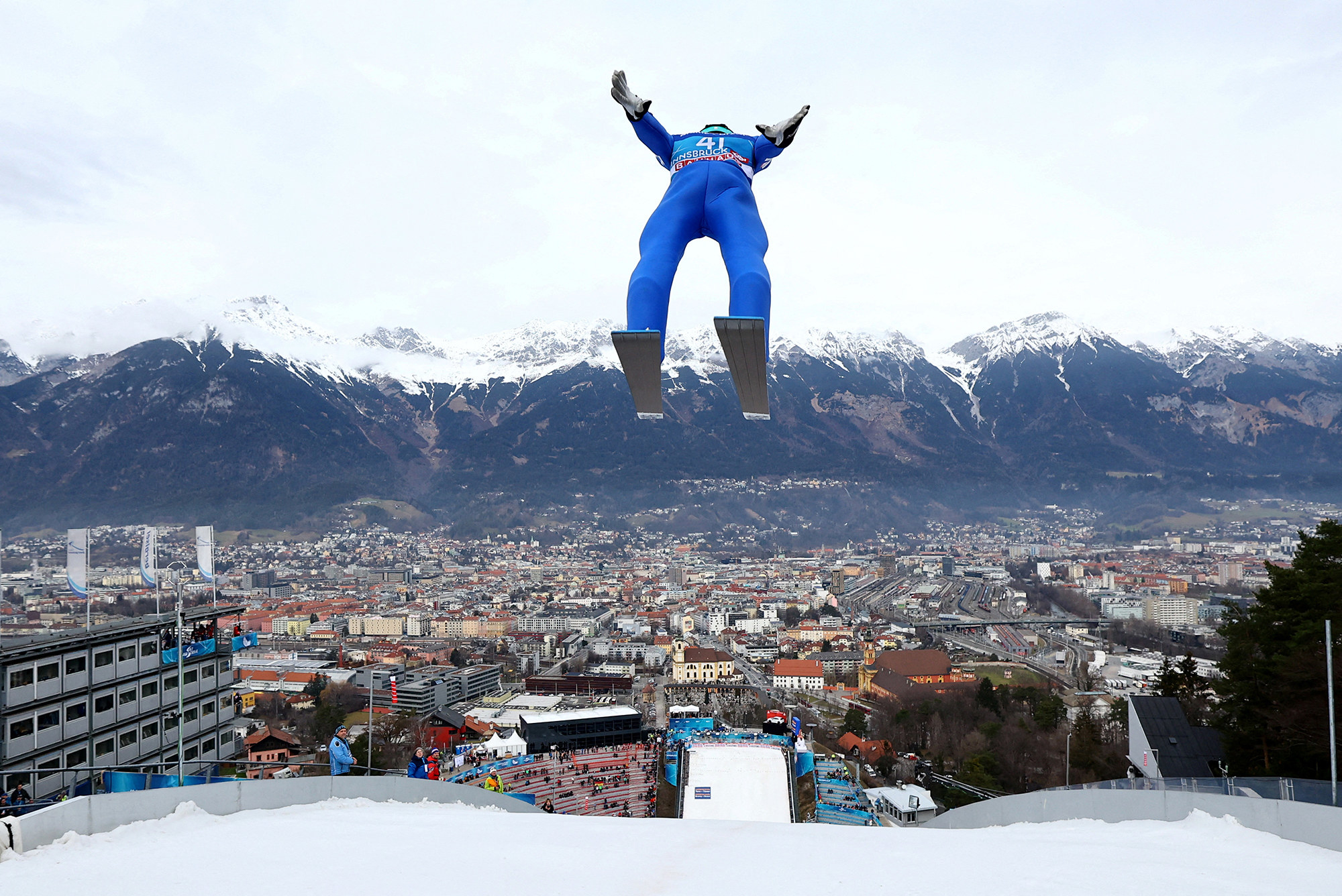 a man in a blue jumpsuit and skis jumps backwards; in the background are snowy mountains and a city