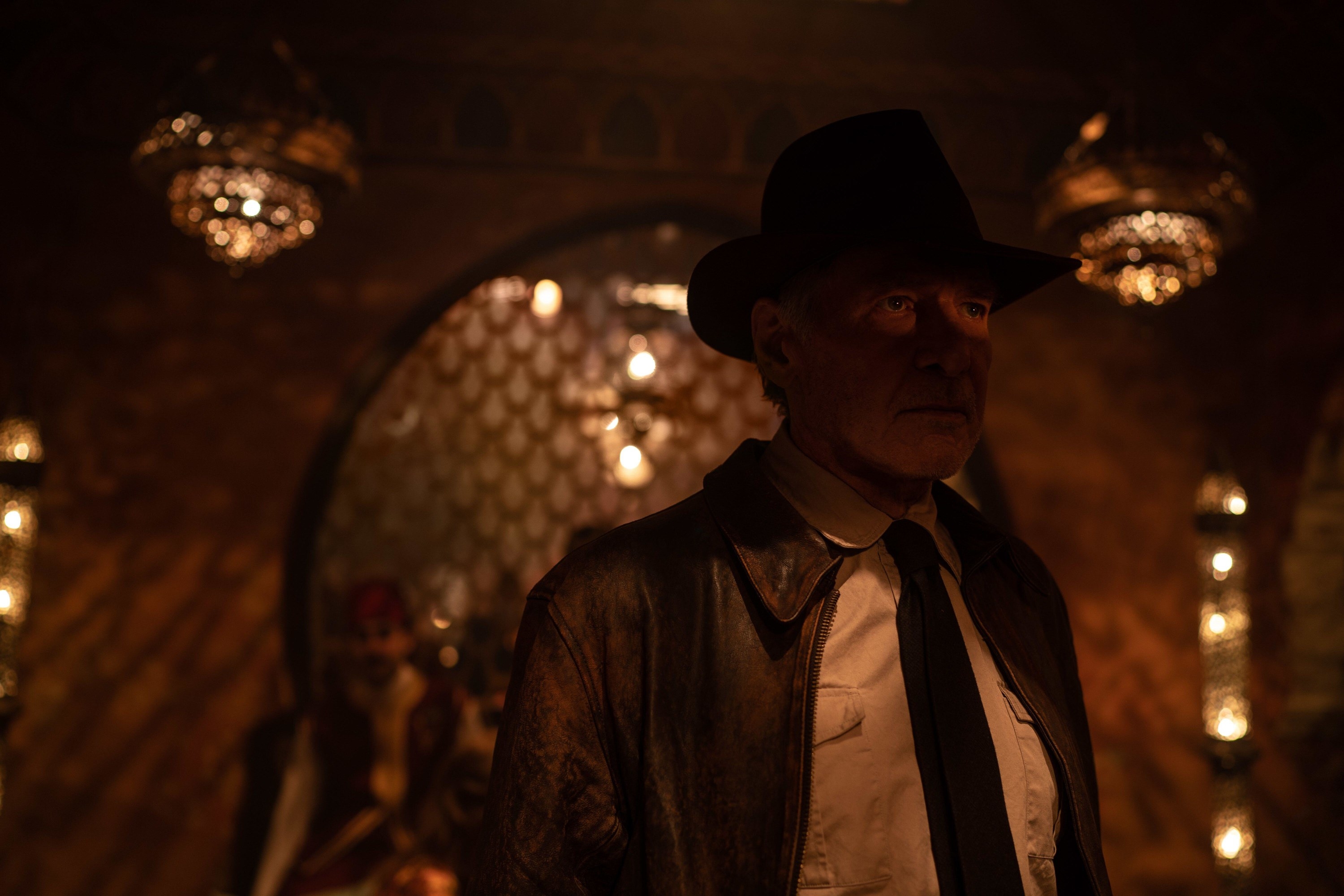 An older man in a hat and leather jacket enters a dimly lit room