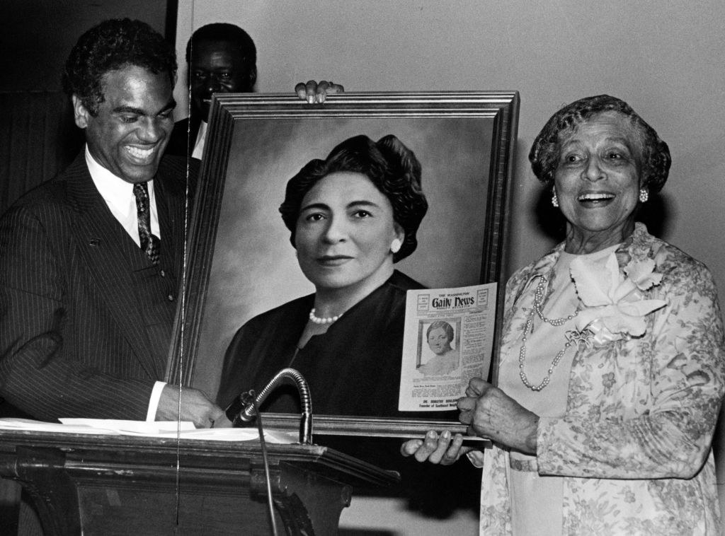an older dr dorothy stands beside a man presenting a portrait of her