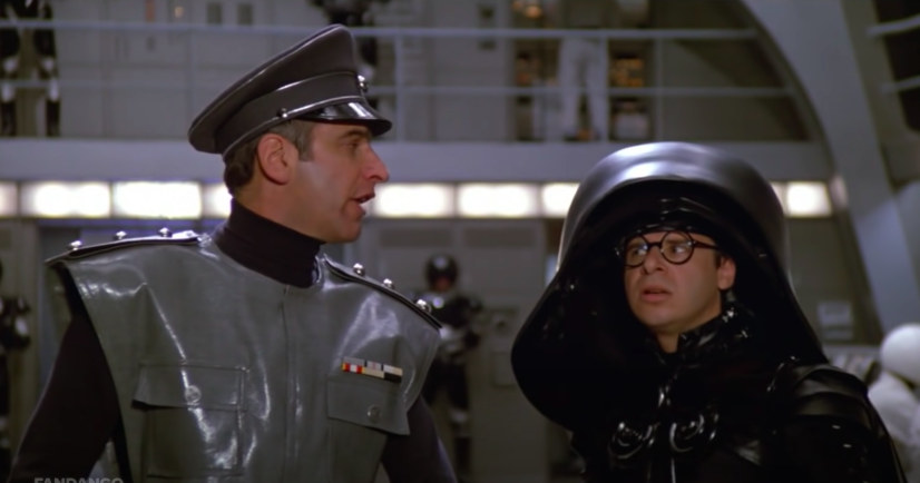 A man in uniform looks at another man wearing a huge helmet