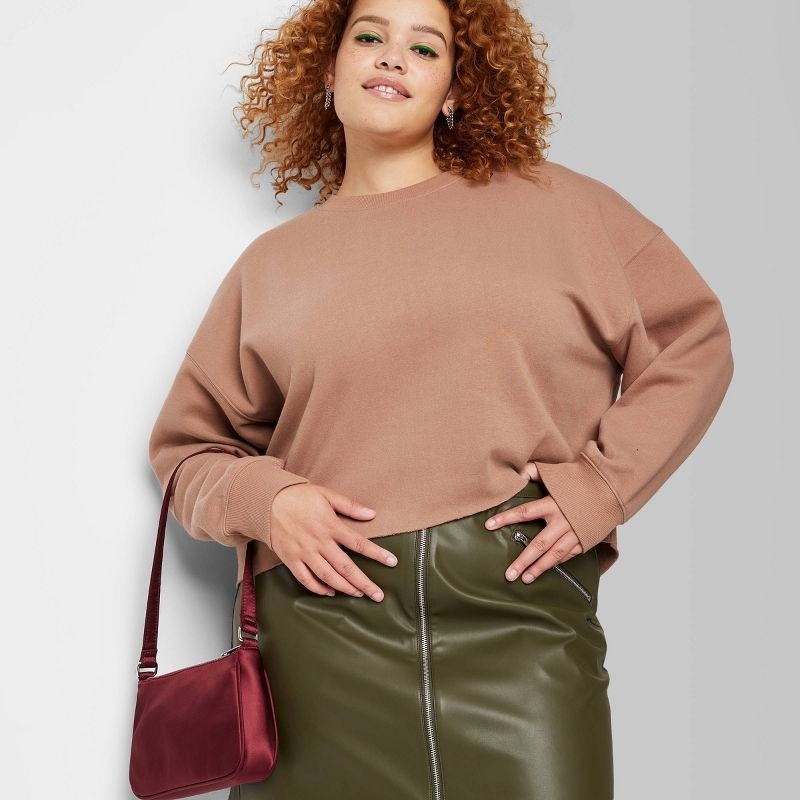 Model wearing brown cropped pullover with green skirt, holding red purse