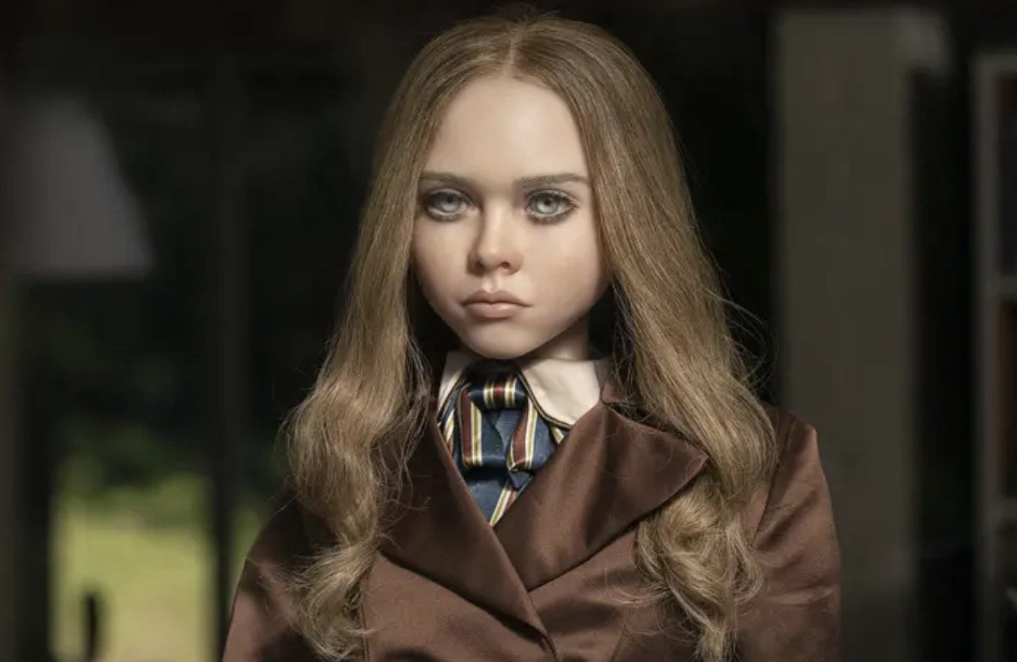 A realistiic ish looking M3GAN doll stares ahead wearing a brown suit
