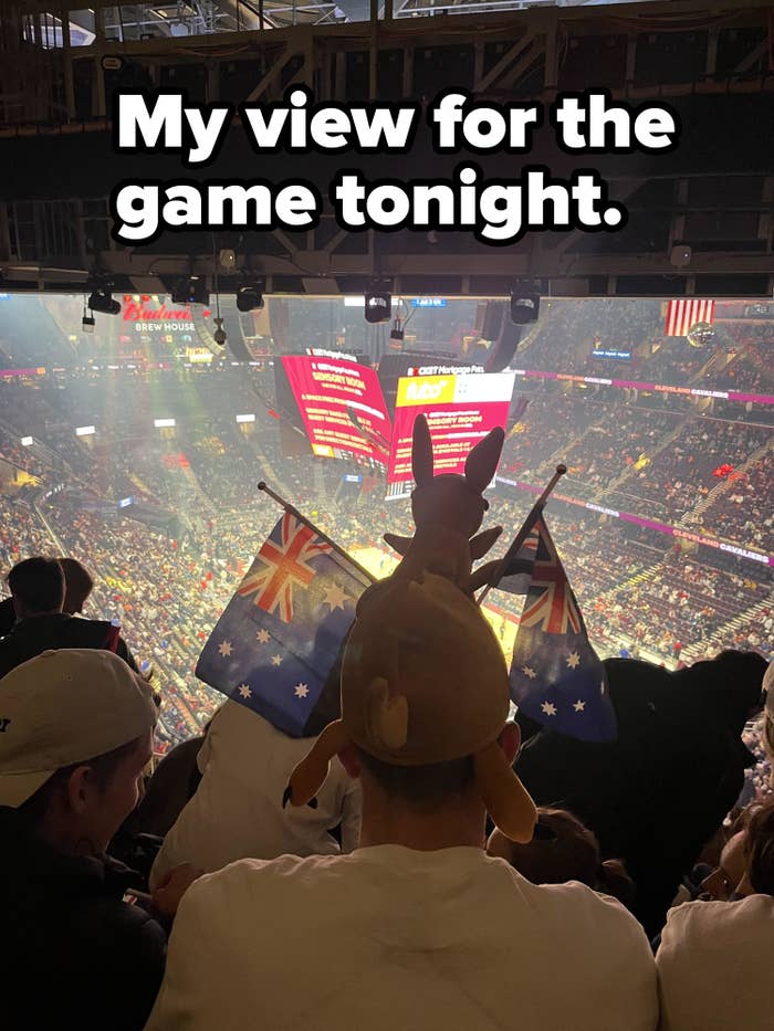 Person at a sports event with someone wearing a kangaroo hat and holding up two Australian flags and blocking their view