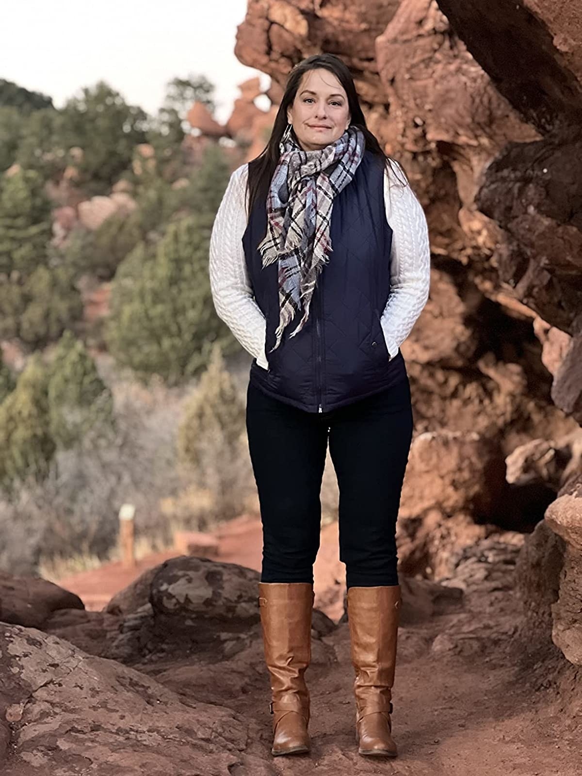 A reviewer in black pants, brown boots, a white top, blue puffer vest and multi-colored scarf