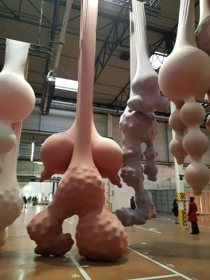 Stretched-out sculptures hanging from the ceiling with large descending balls, some with nipples, at the bottom
