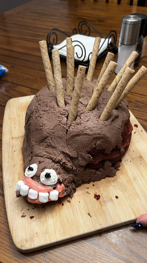 A misshapen, badly frosted cake with a scary face with marshmallow teeth jutting out of one side, and large wafer sticks on top of the cake