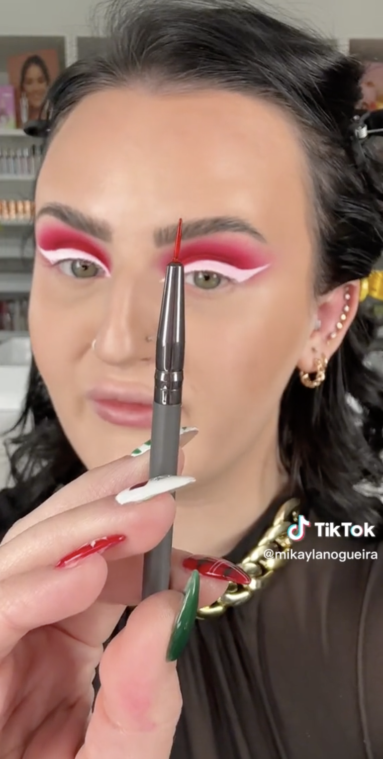 A woman with vivid red and white eyeliner holds a fine makeup brush to the camera