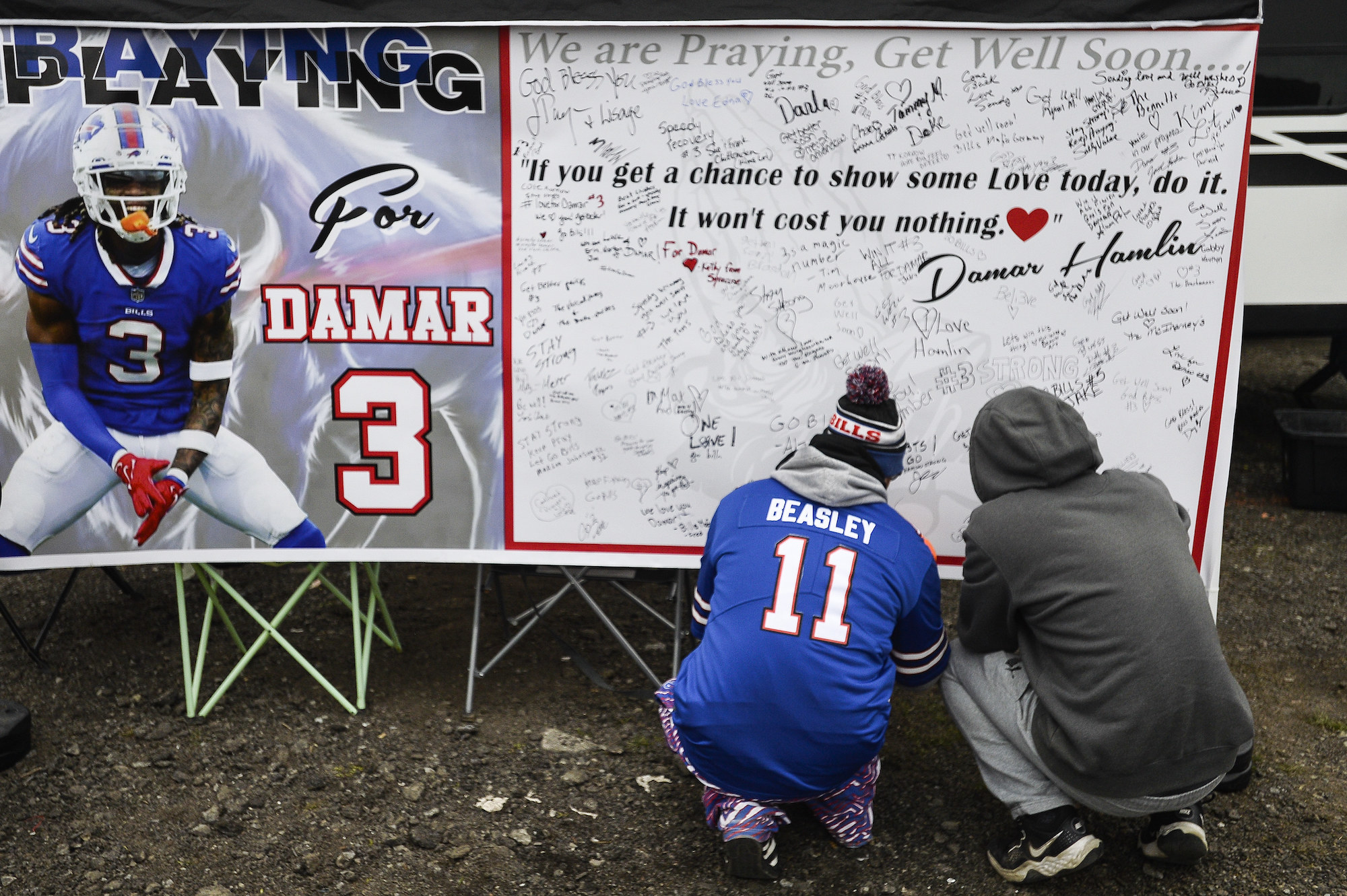 Two fans crouching down to sign a board with &quot;Damar 3&quot; and many handwritten messages on it