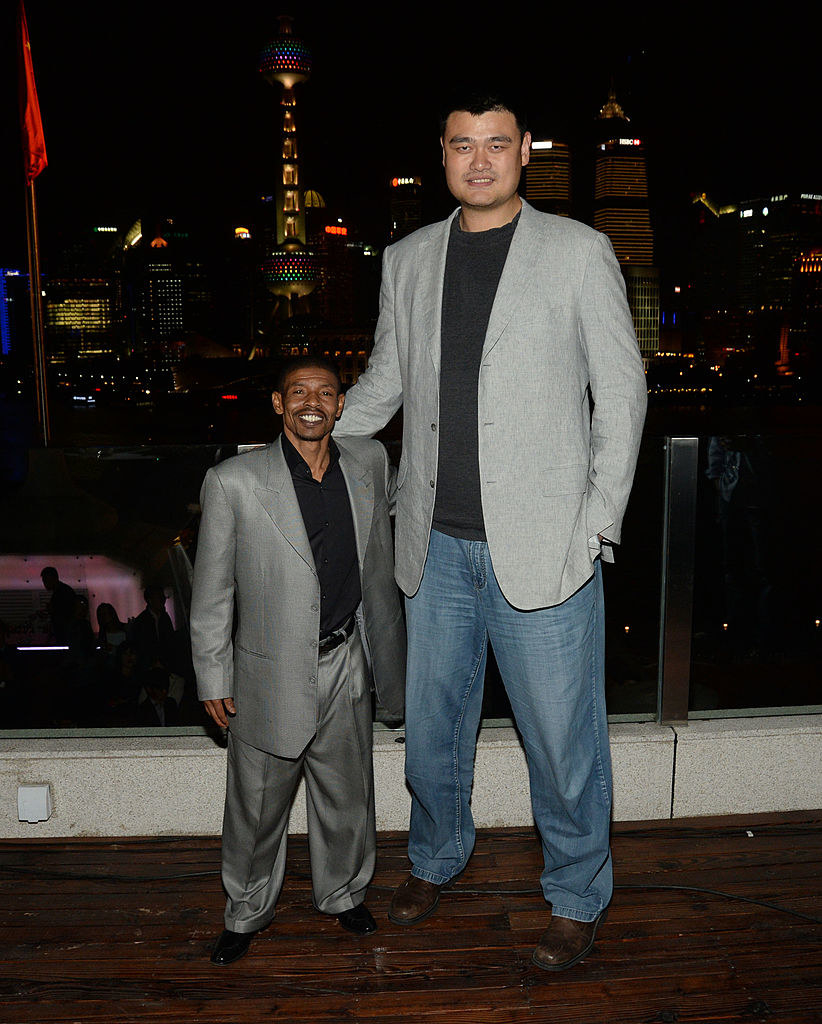 Muggsy, 5 feet 3, in a loose suit and Yao, 7 feet 6, in a suit jacket and loose jeans, standing together in front of a window showing a city skyline