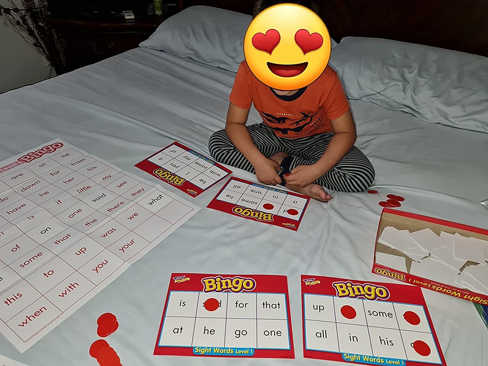 A reviewer&#x27;s child on their bed with the game set up with bingo cards, red chips to mark the words, and a large word list