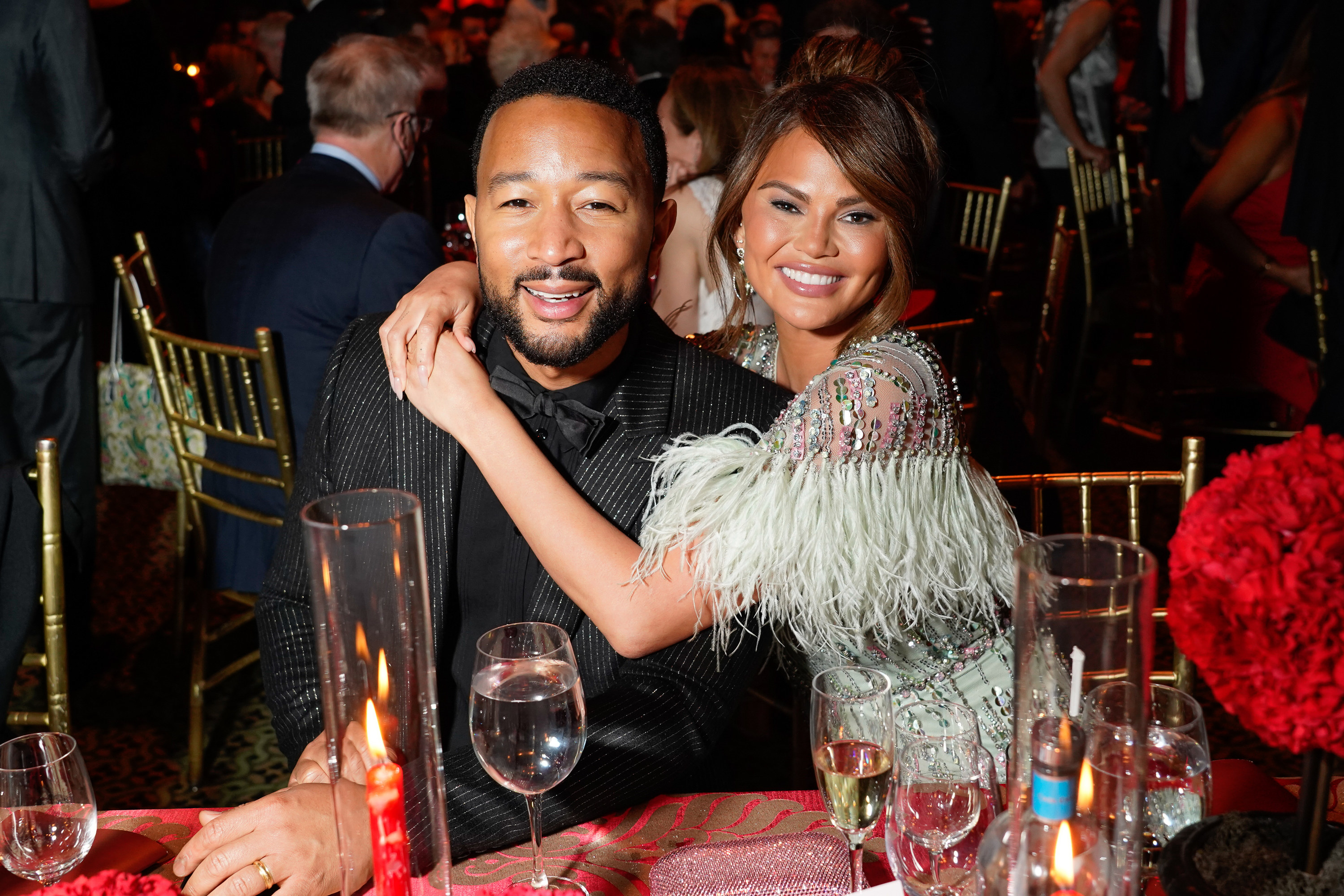 Chrissy has her arms around John as they both smile for a photo while sitting at a table an event