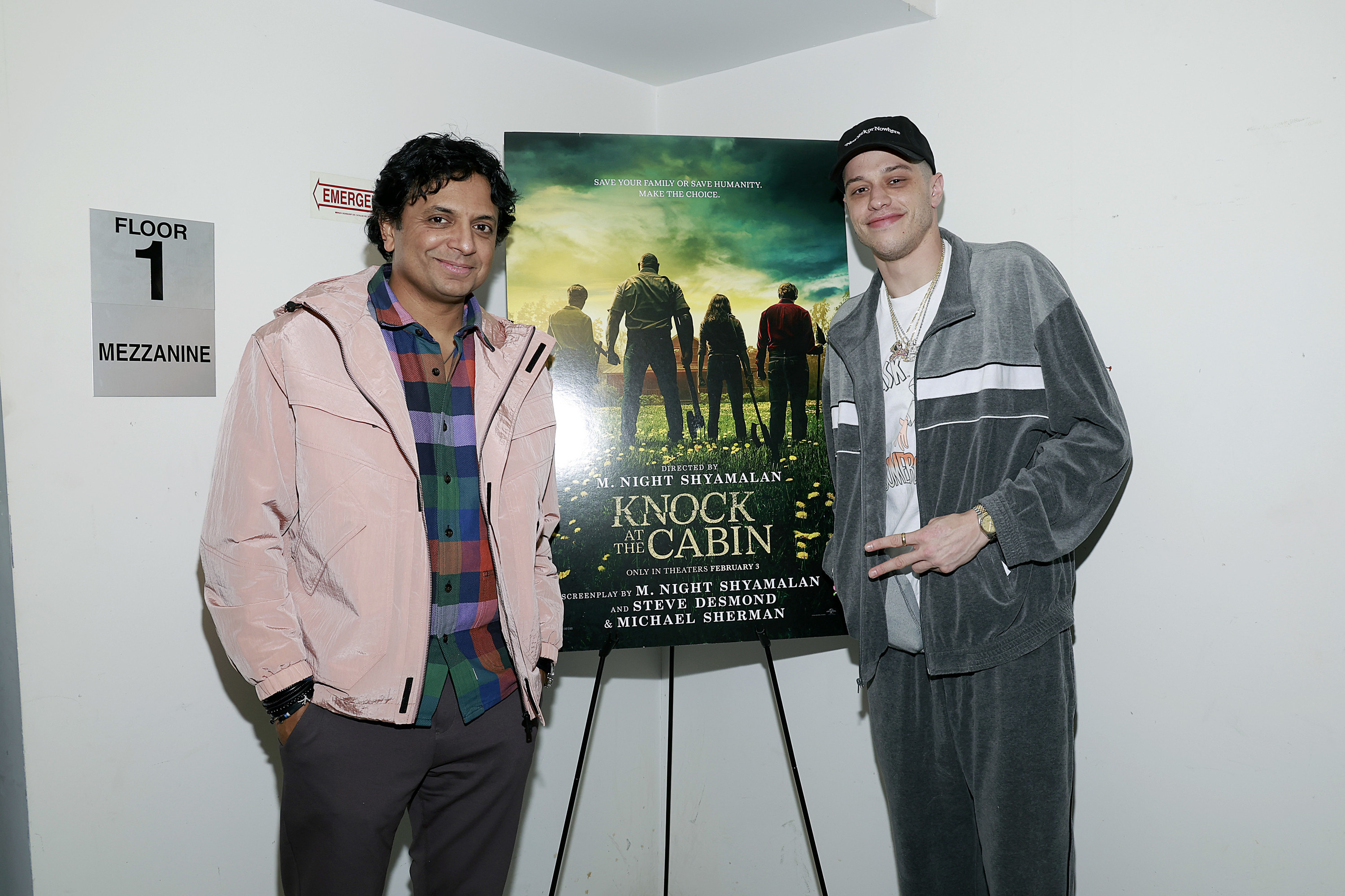 M. Night Shyamalan and Pete stand next to a poster for Shyamalan&#x27;s film &#x27;Knock at the Cabin&#x27;. Pete is wearing a baseball cap