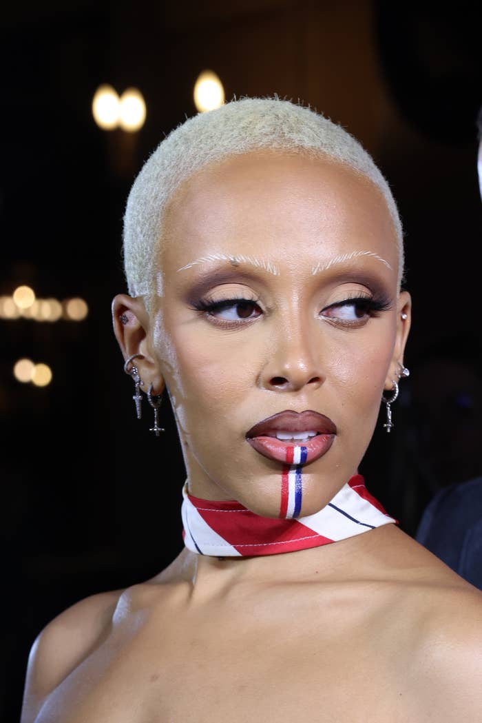 A closeup of Doja at an event rocking a shaved look and tri-colored paint down her bottom lip and chin