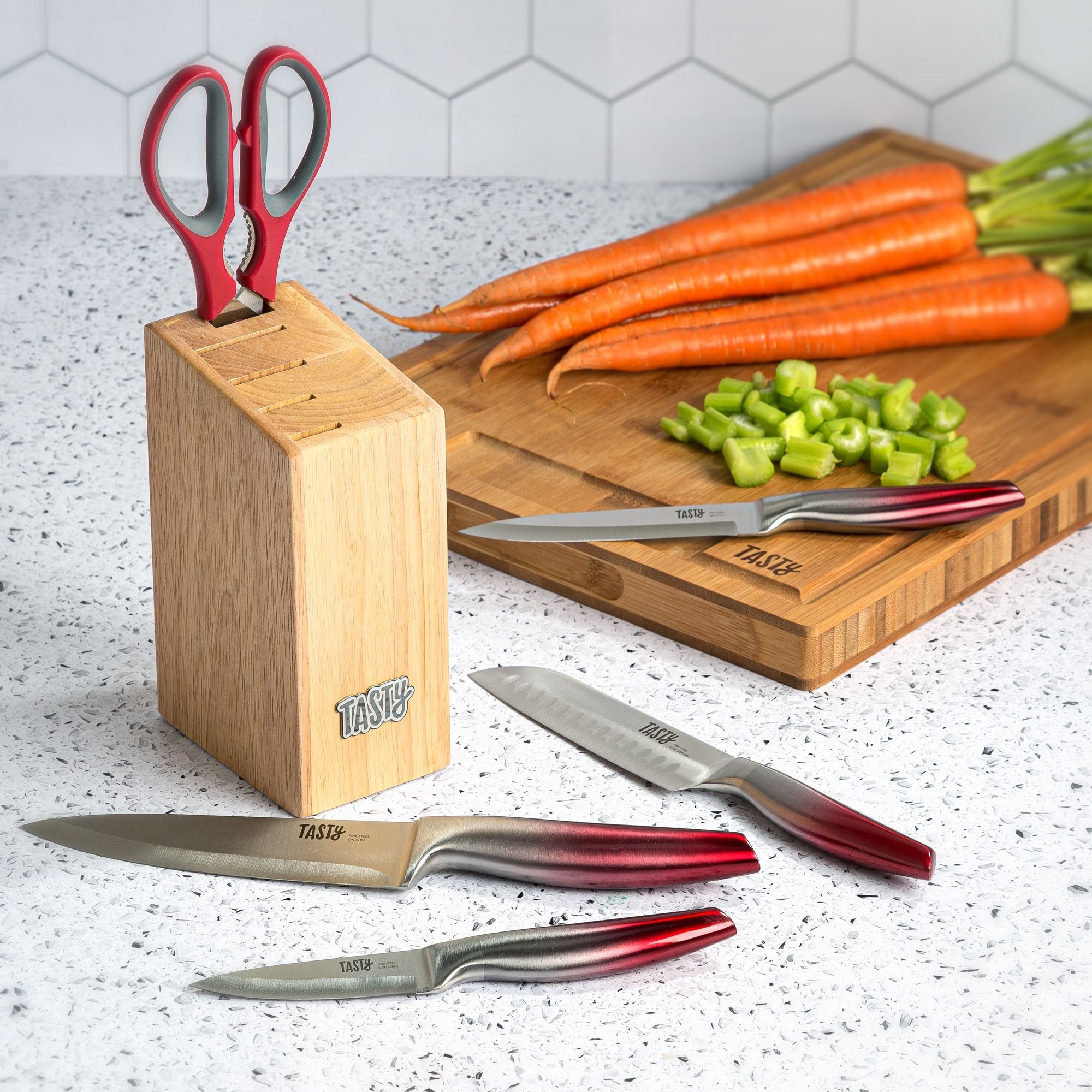 Wooden knife block with red scissors in it, four knives laid out, cutting board with carrots and chopped celery in back