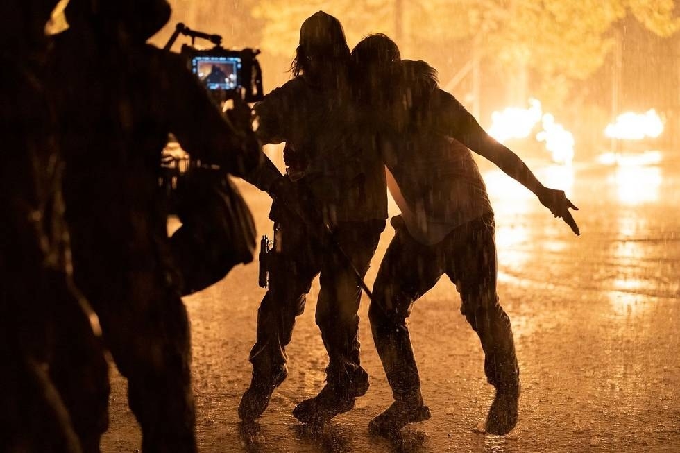 A cameraperson filming a scene with two people walking through the pouring rain with fire behind them