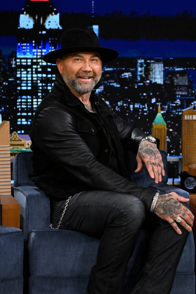 Dave smiles as he sits during a late-night TV talkshow. Dave is wearing a fedora and several pieces of jewelry on his hands