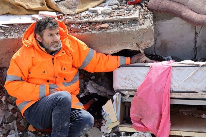 A man in a bright orange jacket sits atop a collapsed building and holds his dead daughter&#x27;s hand
