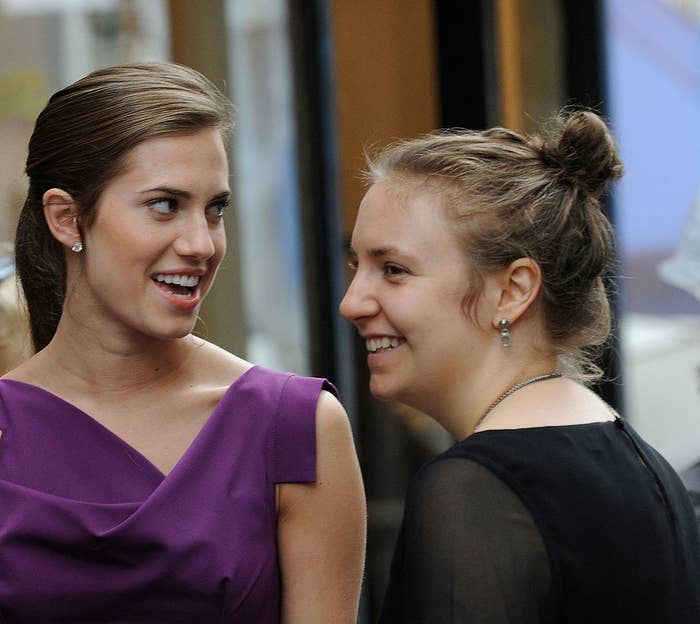 Allison Williams (left) grinning and looking at Lena Dunham (right), who is doing a small smile