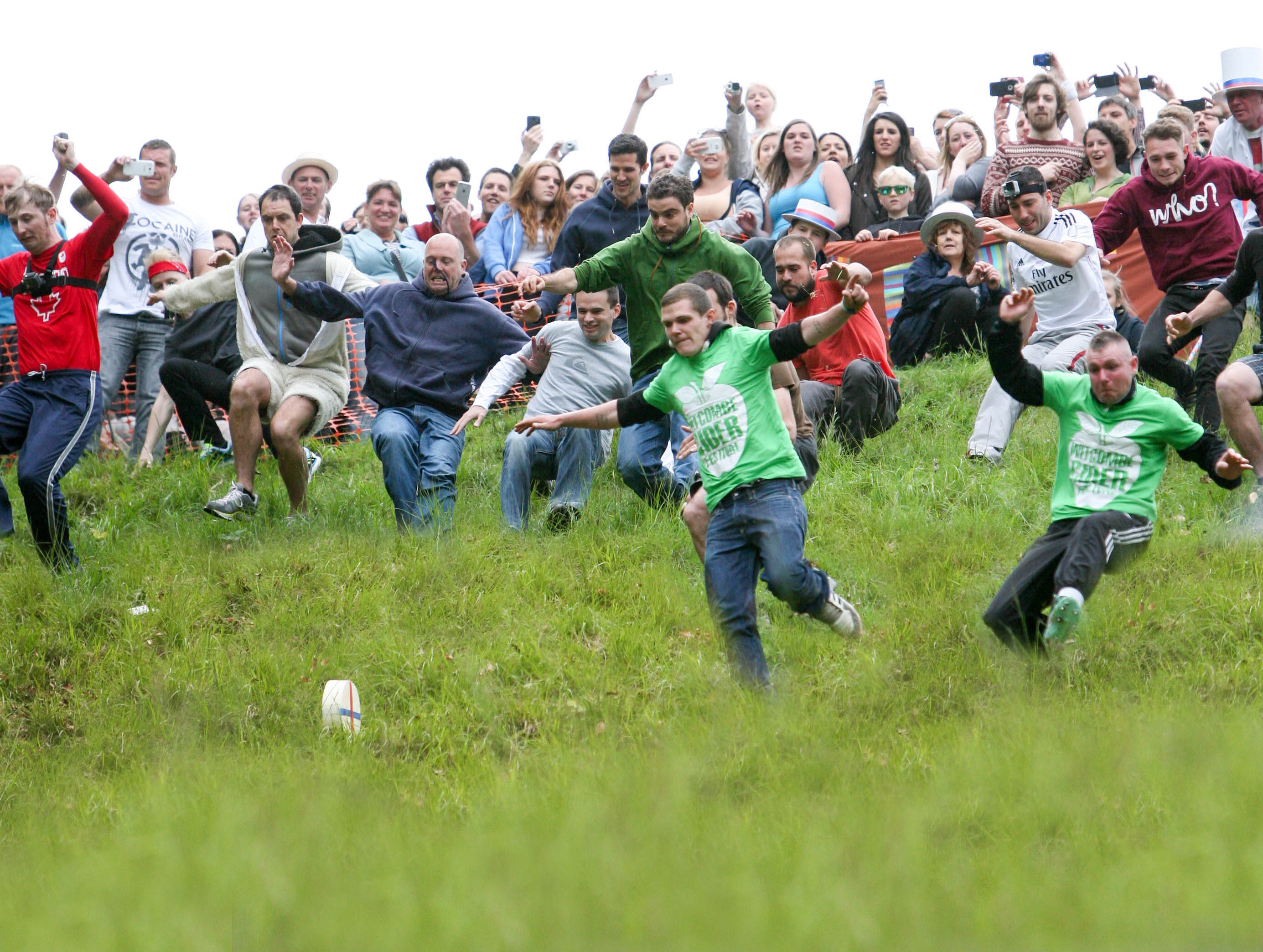 people chase a cheese wheel down a steep hill