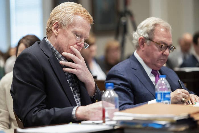 Two men sit with pensive looks, facing a tabletop covered in documents and folders