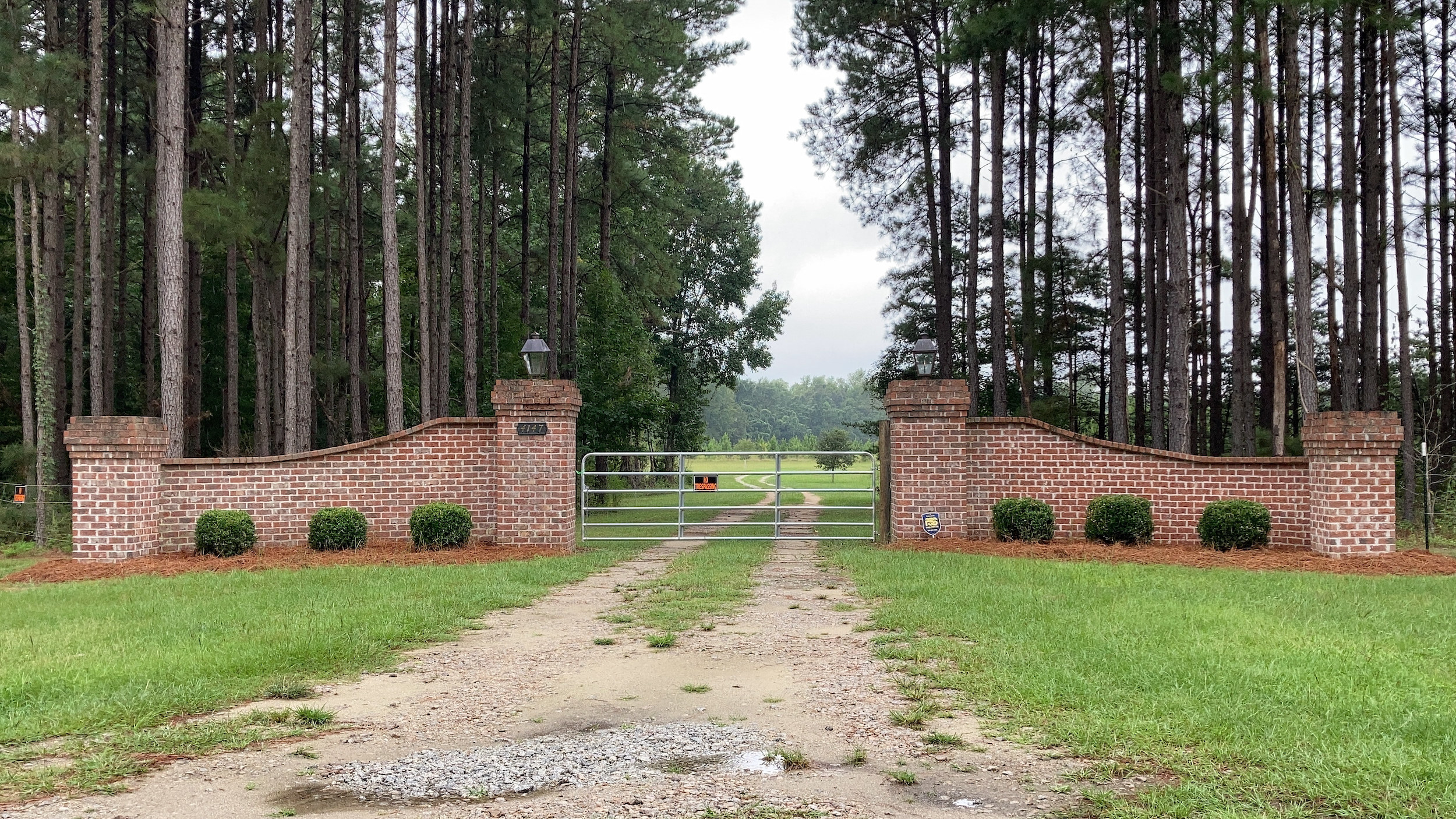 A brick-framed gate with a &quot;no trespassing&quot; sign in front of a road leading into a meadow, surrounded by tall, spindly trees