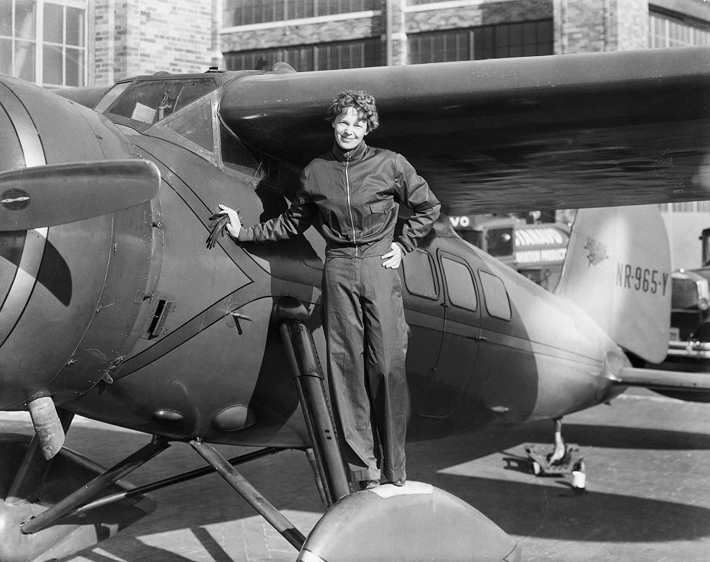 Earhart smiling for a camera while standing on the wheel of a small plane