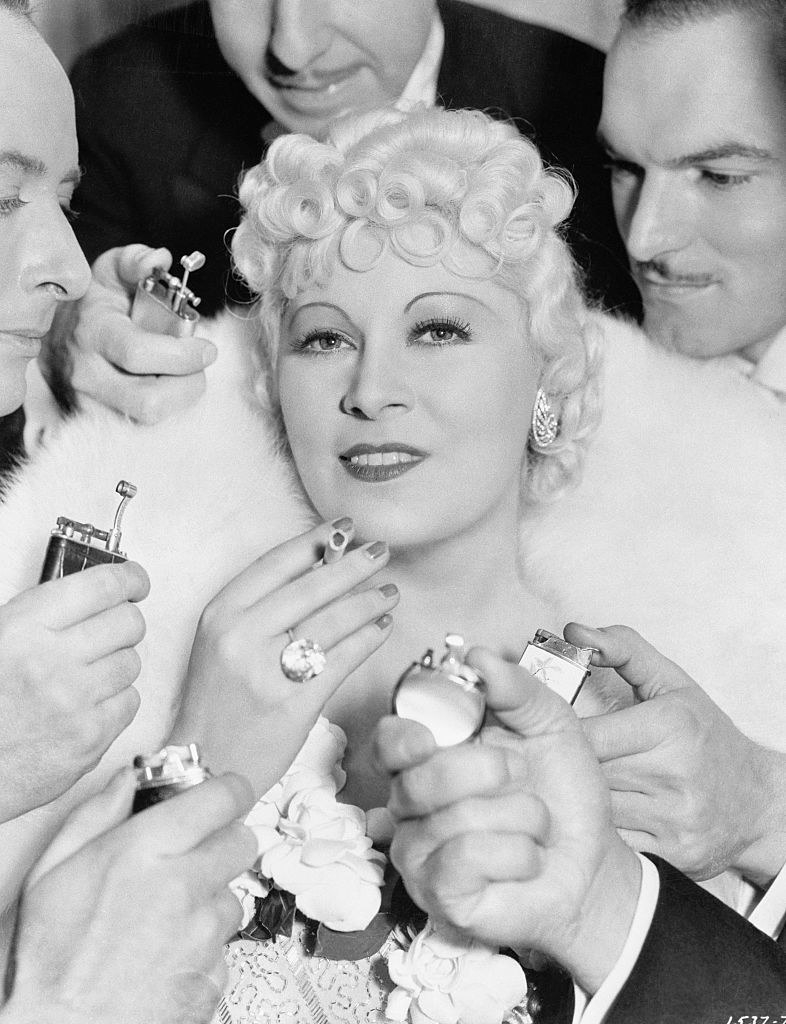 Close-up of Mae holding a cigarette and surrounded by men with cigarette lighters