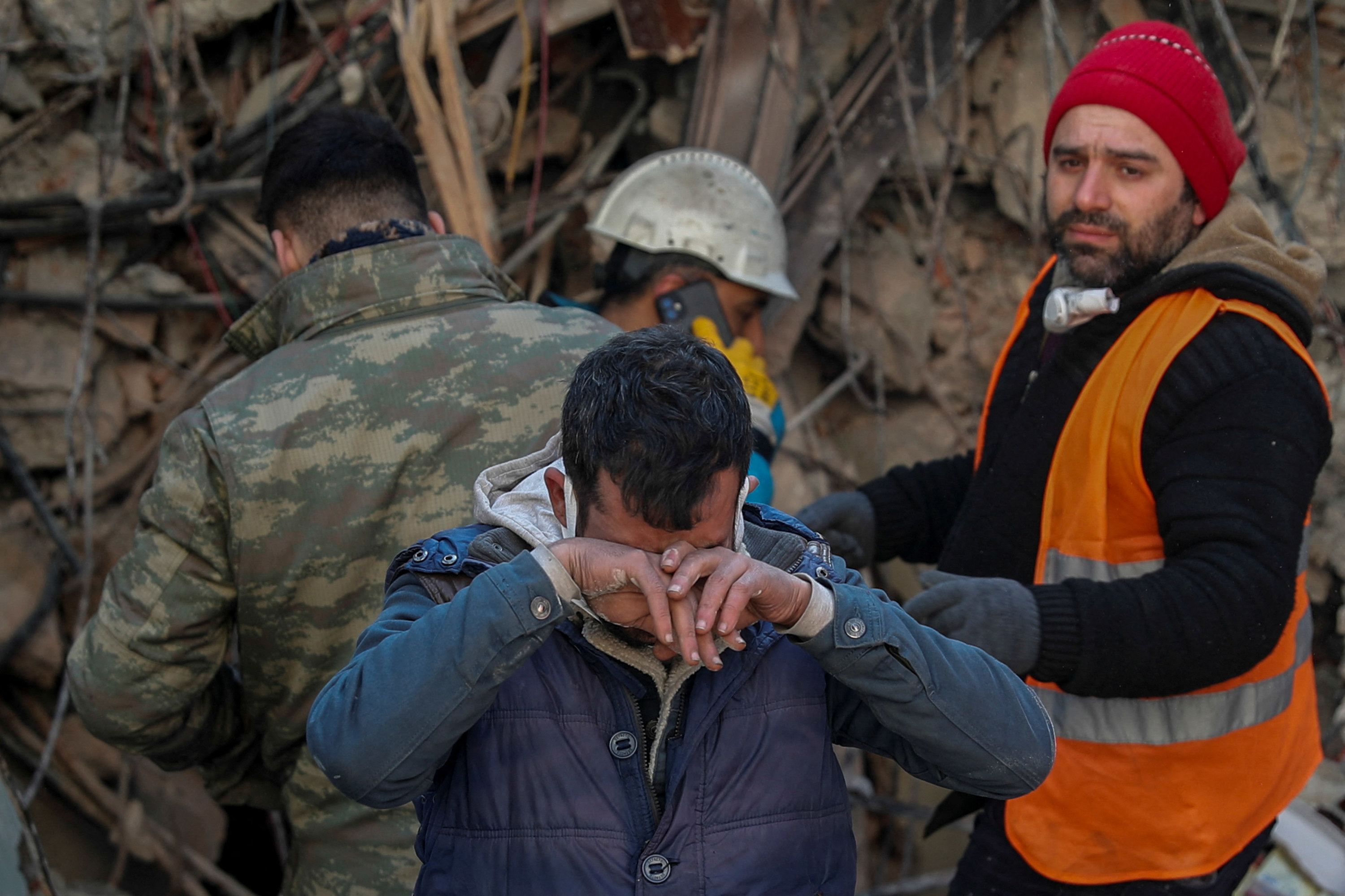 A man covers his eyes as two men stand behind him in front of rubble