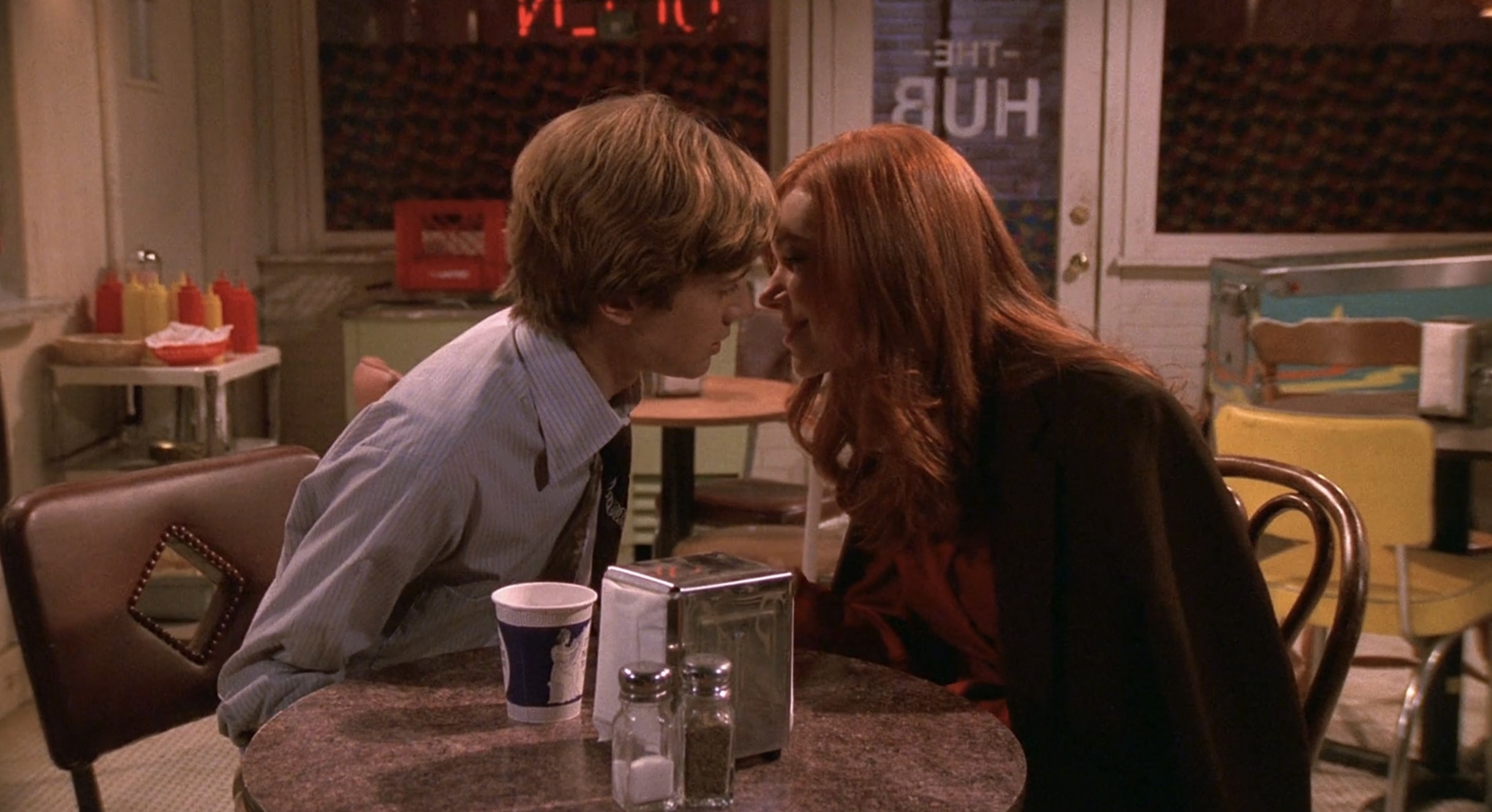 Eric and Donna sit at a table in The Hub and lean in to kiss each other