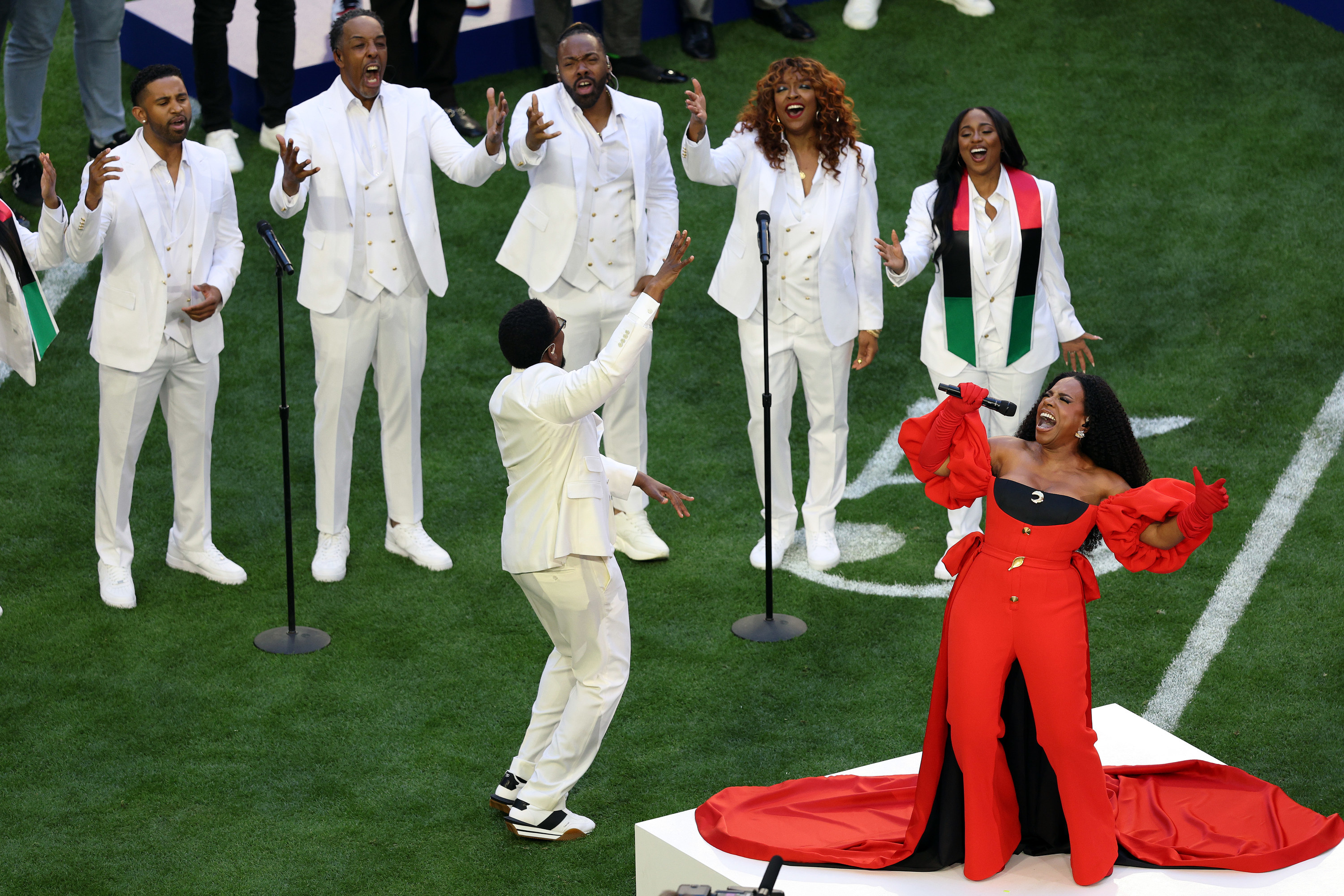 Sheryl performing on a platform on the football field in a pantsuit with flowy sleeves and long train with backup singers behind her