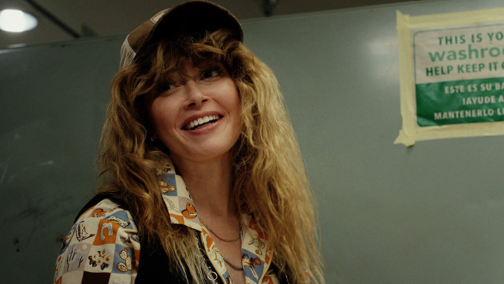 A blonde woman in a trucker hat and vest smiles in a bathroom