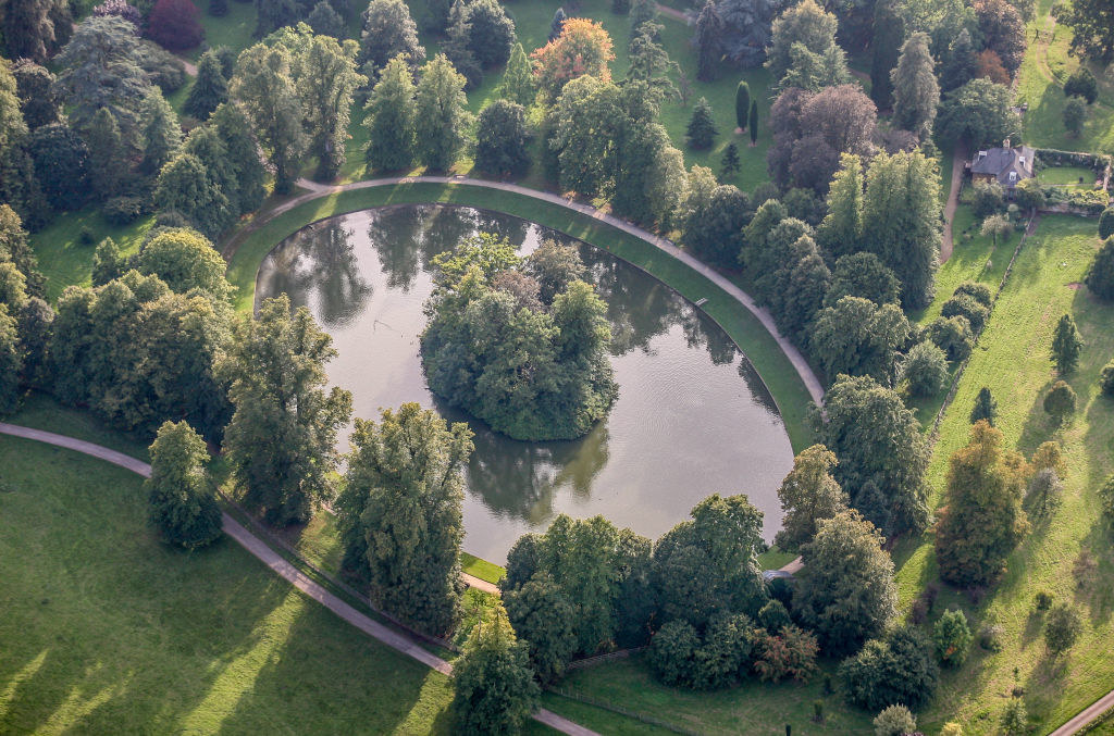 A tiny round piece of land with bushes in the middle of a pond, surrounded by a large garden
