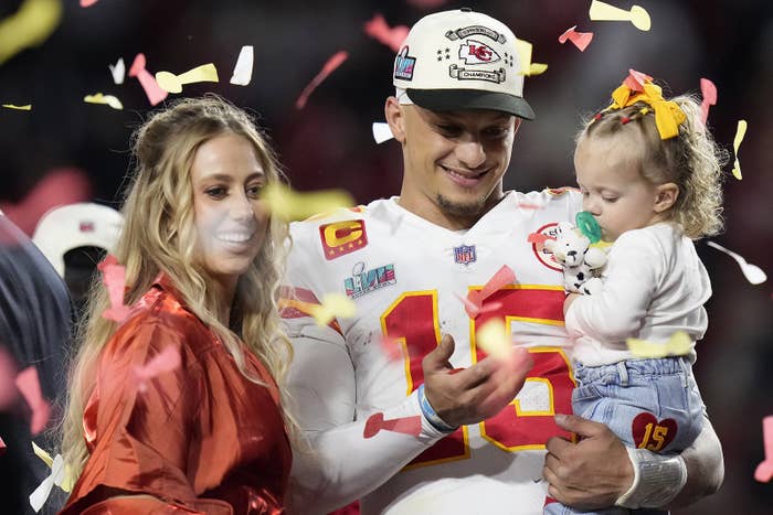 A smiling quarterback holds a toddler in his arms, standing beside his smiling wife as confetti falls around them