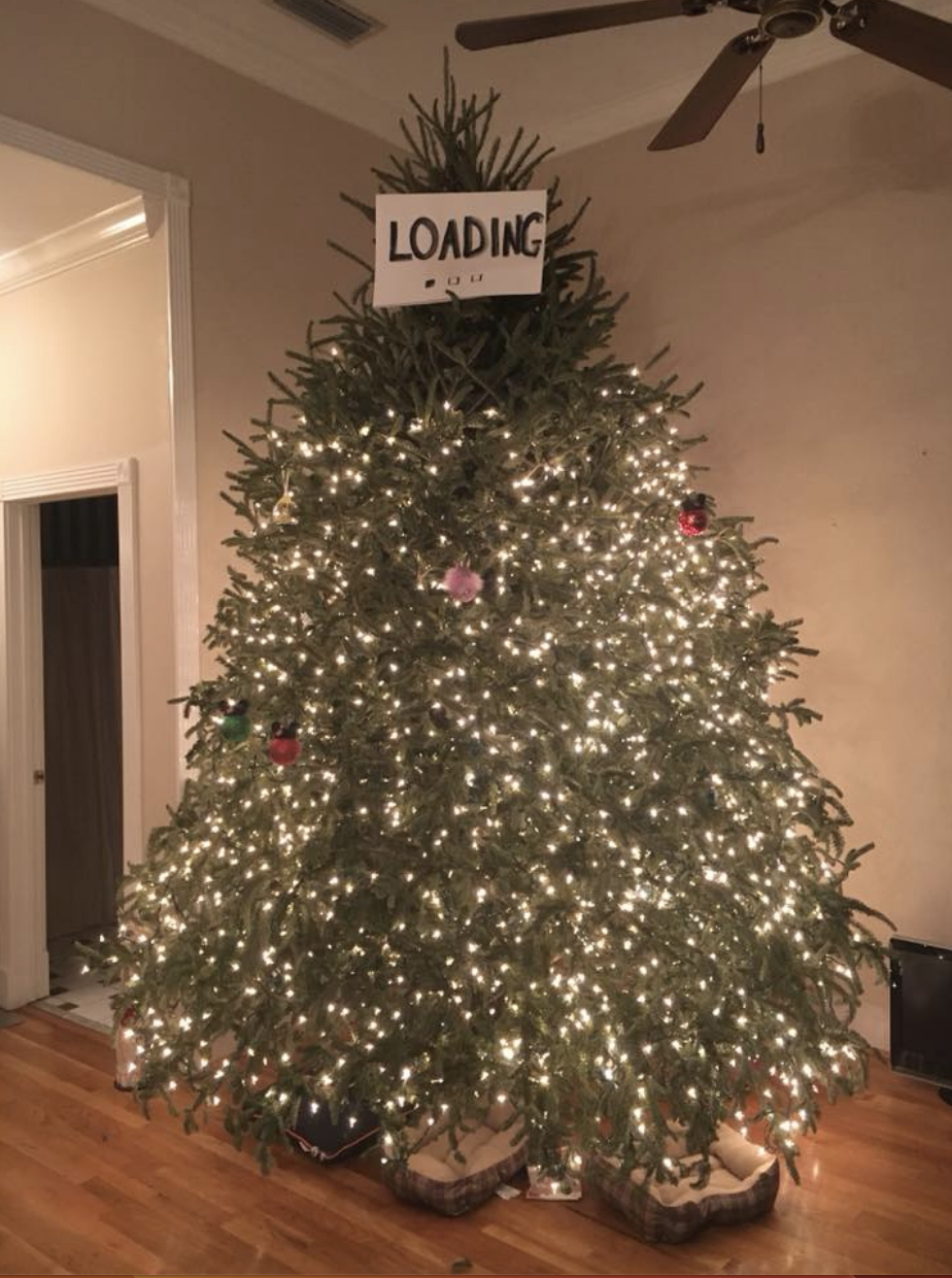 A Christmas tree is covered in lights until about 80% of the way up, where the lights stop and there is no tree topper, only a paper sign saying &quot;loading&quot;