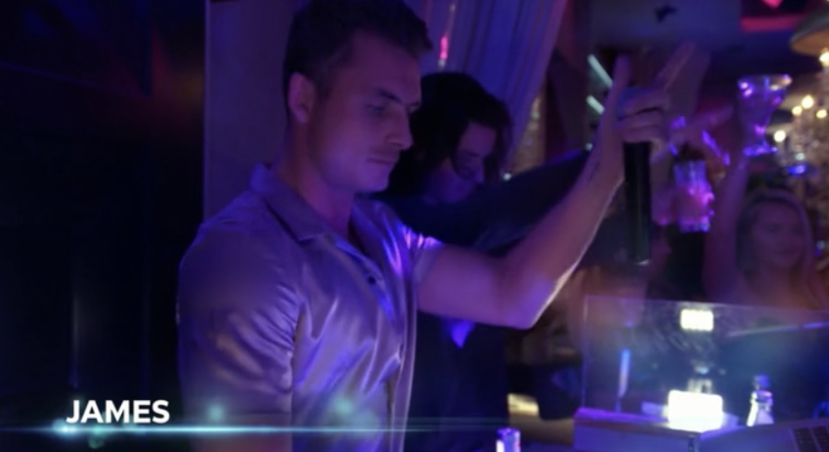 James stands behind a DJ booth with a microphone in a satin purple shirt