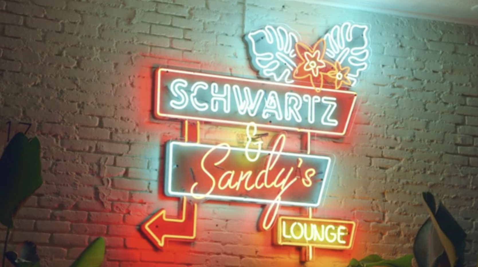 An orange and teal neon sign mounted on a white brick wall, it reads Schwartz &amp;amp; Sandy&#x27;s Lounge