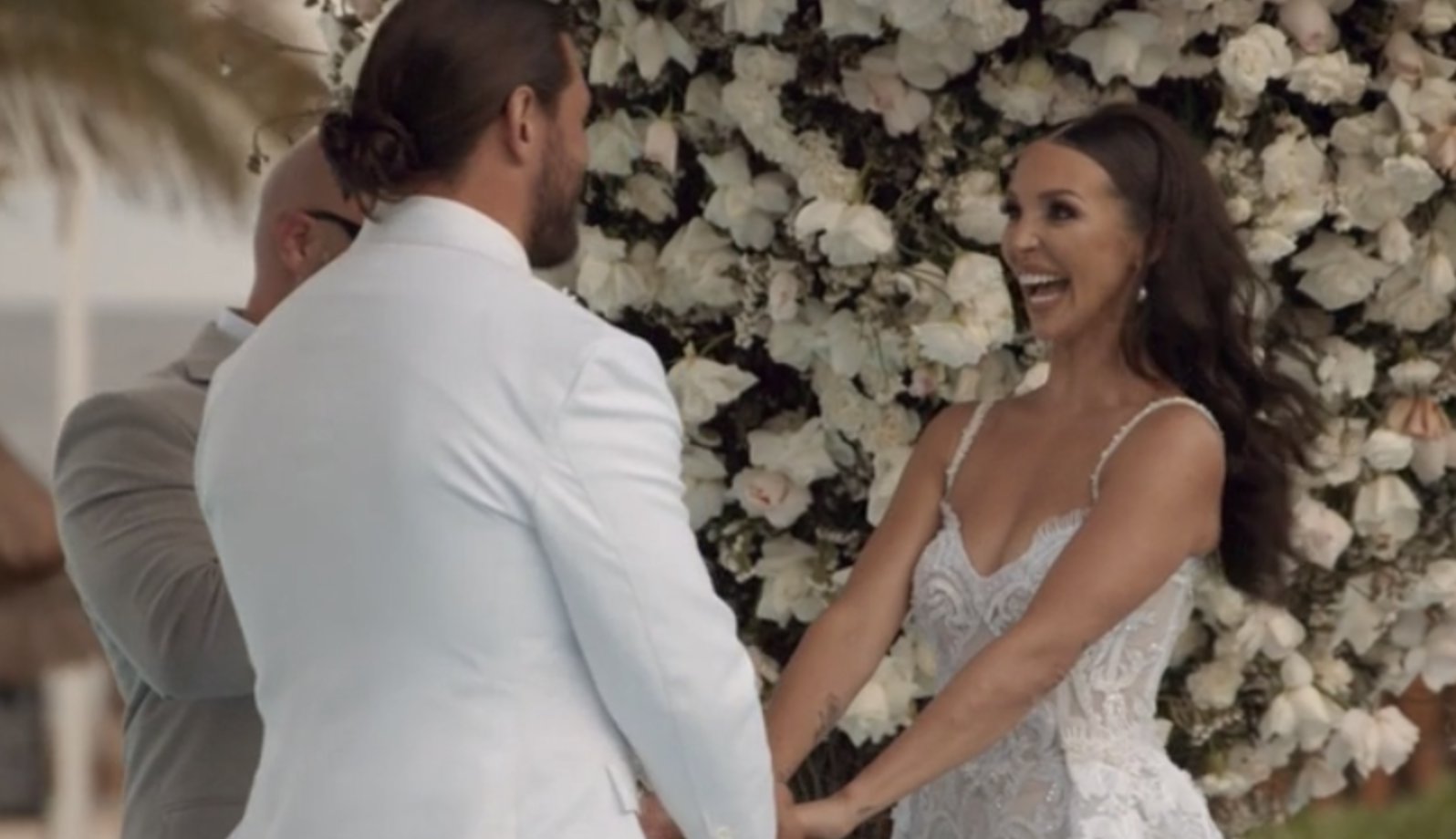Scheana is standing at the altar in a white dress, with a wall of white roses behind her, looking INCREDIBLY EXCITED and holding hands with Brock who is wearing a white suit
