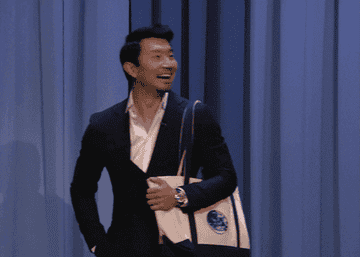 Simu Liu walking onstage smiling big and raising hand to wave with tote bag slung over his shoulder