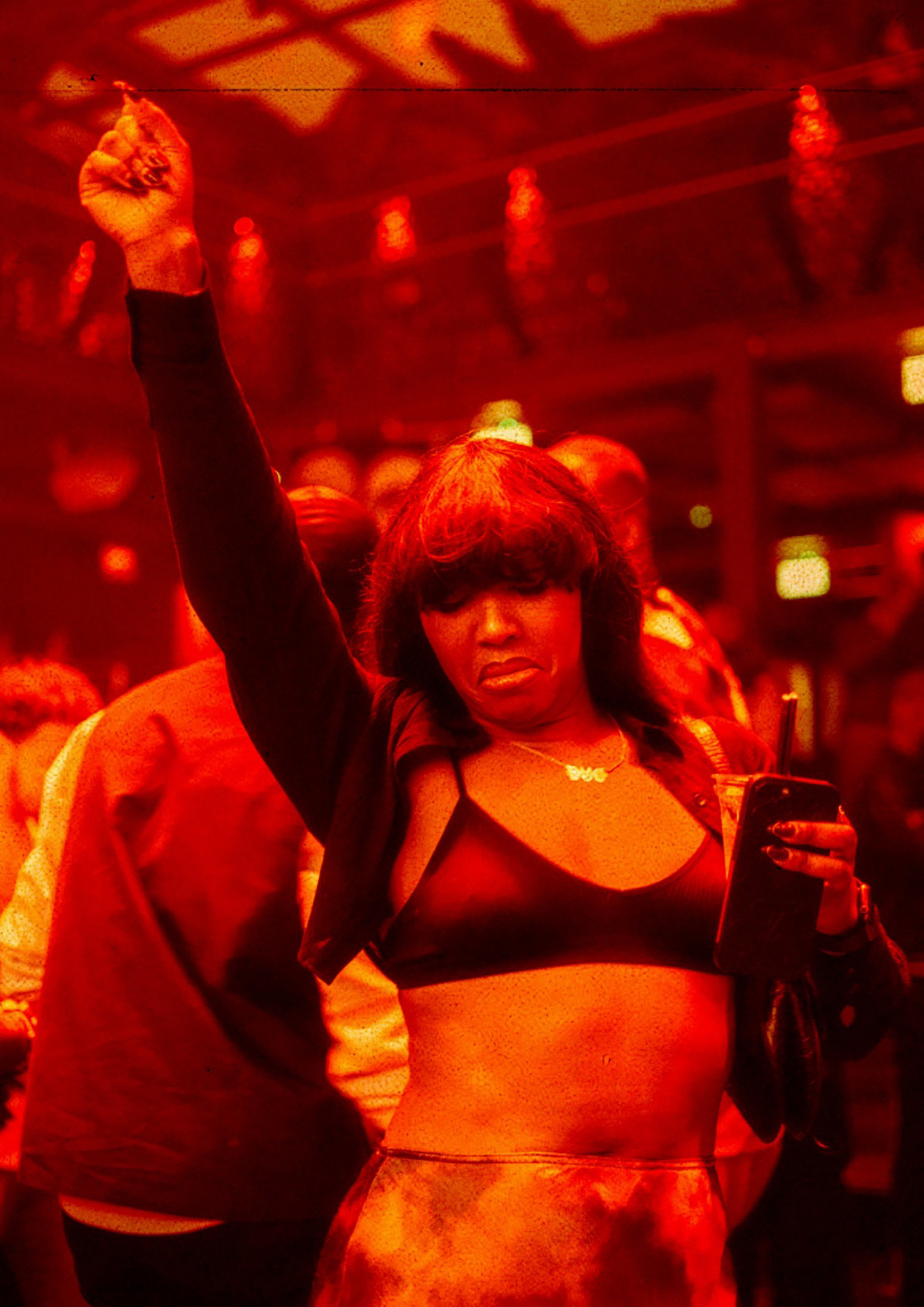 A Black woman dances in a club while holding her drink and a mobile phone.